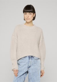 Frau mit kurzen dunklen Haaren, die einen beigen grob gestrickten Pullover und hellblaue hoch taillierte Jeans trägt, steht vor einem einfarbigen grauen Hintergrund.