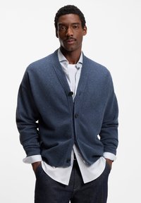 Blå stickad kofta med sjalkrage, knäppning och ribbade muddar, bärs över en vit skjorta, kombinerad med mörka jeans.