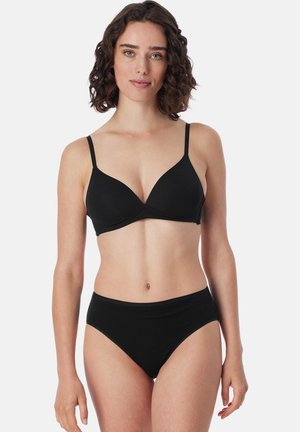 Kobieta o kręconych włosach, ubrana w czarny biustonosz i dopasowane majtki z wysokim stanem, stojąca na tle jasnego, jednolitego tła.