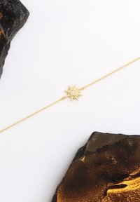 Bracciale d'oro con un ciondolo a forma di sole con una pietra centrale, posato su una delicata catena contro rocce scure e texture.