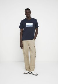 Μπλε βαμβακερό μπλουζάκι με ορθογώνιο λογότυπο σε άσπρο και μπλε, συνδυασμένο με χακί παντελόνι cargo με πολλές τσέπες και μανσέτες.