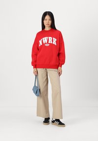 Felpa rossa con testo bianco "NWRK", pantaloni beige a gamba larga e sneakers neri. Tiene una piccola borsa blu. Design semplice e vestibilità casual.