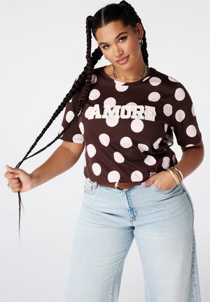 Femme aux longues tresses portant une chemise marron à pois affichant "AMORE" et un jean bleu clair, tenant une tresse et posant avec confiance.