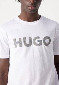 Hvit bomulls T-skjorte med en stor "HUGO" logo. Teksten har et strukturert, rutete mønster med grå og blå aksenter.