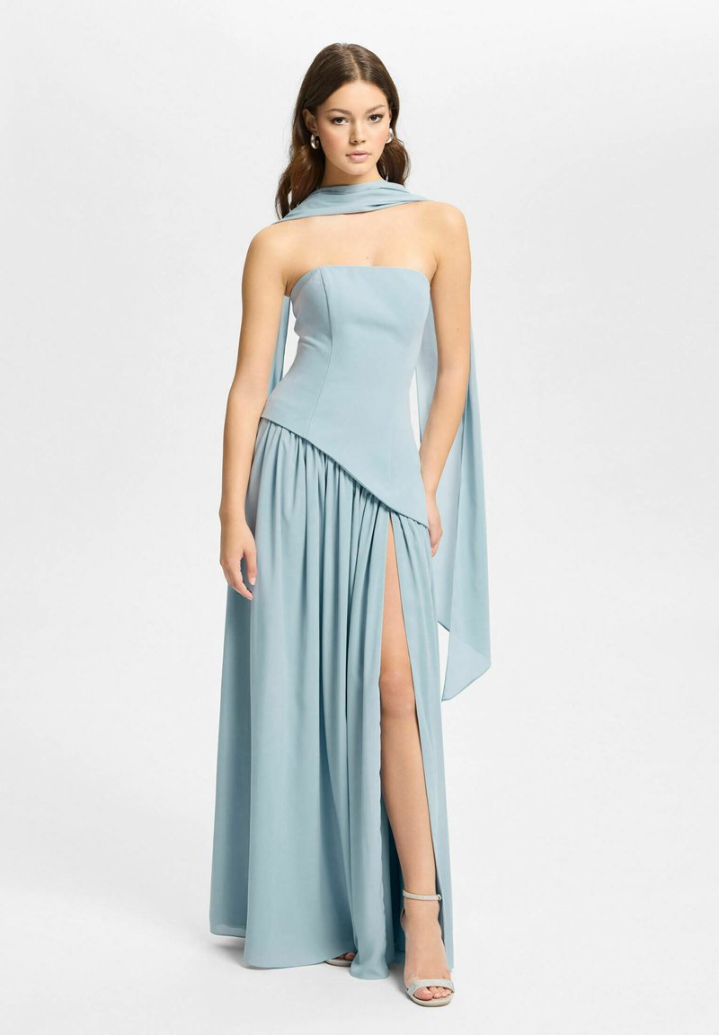 Femme portant une robe bustier bleu pâle avec un corsage asymétrique, une fente haute jusqu'à la cuisse, une écharpe assortie et des talons blancs à bride à la cheville sur un fond uni.