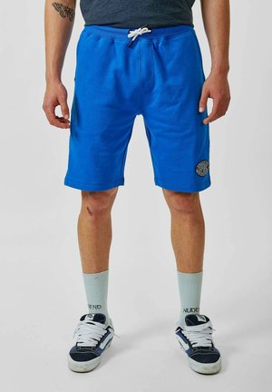 Shorts en coton bleu avec une taille élastique et un cordon de serrage. Présente une étiquette de logo sur la cuisse gauche. Portés avec des chaussettes et des chaussures bleu clair.