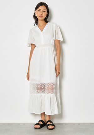 Kobieta ubrana w białą sukienkę midi z koronkową wstawką i falbanowymi rękawami, w czarnych sandałach na odkryte palce, stojąca na tle gładkiej ściany.