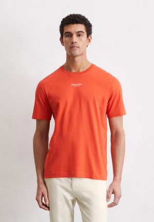 Oranges Baumwoll-T-Shirt mit Rundhalsausschnitt, kurzen Ärmeln und einem weißen Logo, das auf der Brust gedruckt ist. Kombiniert mit hellen Hosen.