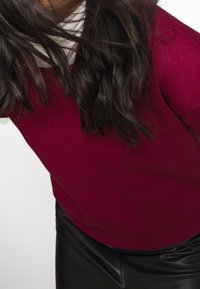 Suéter de punto burdeos con escote redondo, combinado con pantalones de cuero sintético negros. Su largo cabello oscuro y ondulado enmarca el cuello.