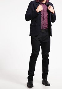 Veste en mélange de laine marine avec col à pression, devant boutonné et poches, associée à une chemise à carreaux bordeaux et un pantalon slim noir, portée avec des bottes noires.