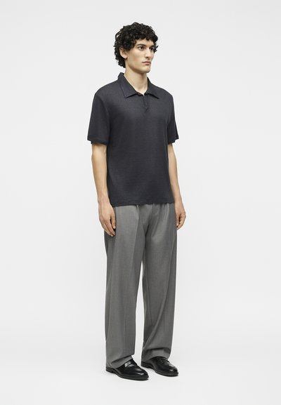 Άνδρας με σγουρά μαλλιά που φοράει μαύρο πόλο μπλουζάκι με κοντά μανίκια, γκρι παντελόνι φαρδιάς γραμμής και μαύρα μοκασίνια, στέκεται μπροστά σε μια μονόχρωμη επιφάνεια.