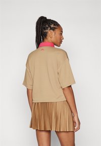 Καφέ κοντομάνικο μπλουζάκι με ροζ γιακά και λογότυπο, συνδυασμένο με πλισέ καφέ φούστα. Το ύφασμα έχει υφή και μοτίβο.
