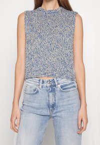Dzianinowy top bez rękawów w kolorze niebieskim i beżowym, z wysokim kołnierzem i krótkim krojem. Połączony z dżinsami w jasnym odcieniu.