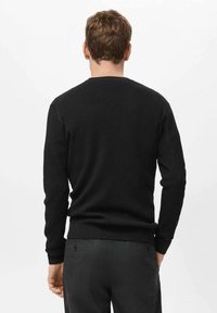 Suéter negro de canalé con mangas largas, cuello redondo y puños ajustados; lleva encima de pantalones gris oscuro, visibles desde la parte trasera.