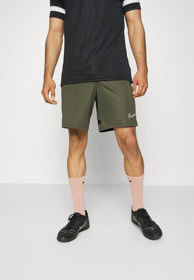 Homme debout portant un t-shirt noir, un short Nike vert olive, des chaussettes Nike rose clair et des baskets noires sur fond uni.