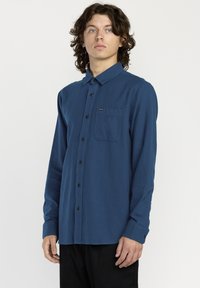 Chemise bleu à manches longues avec une poche poitrine, boutons noirs et une texture douce. Stylée avec un col et une coupe droite.