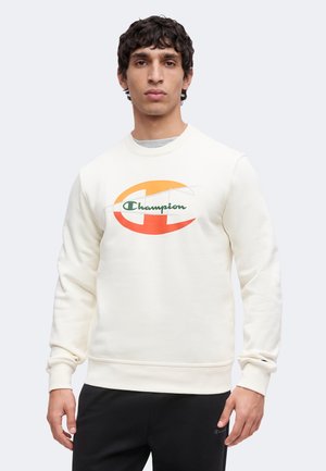 Homme portant un sweat-shirt blanc avec un grand logo Champion orange et jaune, associé à un pantalon noir, debout sur fond blanc.