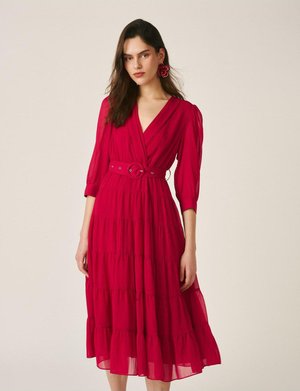 Mujer con un vestido rojo hasta media pierna, mangas abullonadas de tres cuartos, escote en V, cinturón ceñido a la cintura y pendientes de flor roja a juego.