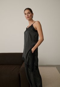 Femme aux cheveux attachés portant une robe noire à pois avec bordure en dentelle, debout à côté d'un canapé brun foncé dans une pièce neutre.
