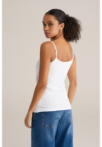 Fehér pamut ujjatlan póló állítható spagetti pántokkal. A háta kerek kivágású. Magas derekú kék farmernadrággal párosítva.