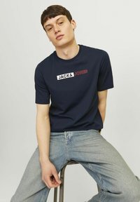 Marineblauwe katoenen t-shirt met een logo ontwerp waarop "JACK&" in het wit en "JONES" in het rood staat. Ronde hals en korte mouwen. Draagt het met lichte spijkerbroek.