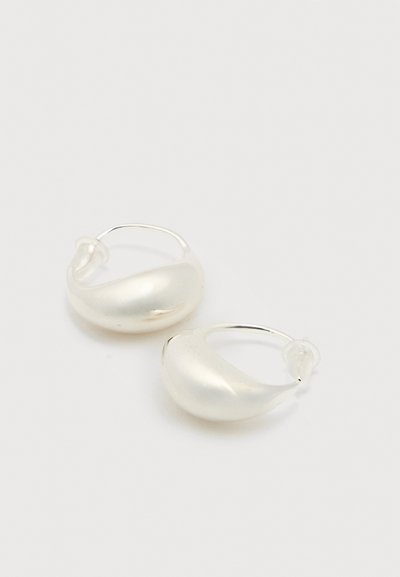 Ζεύγος λαμπερών ασημένιων σκουλαρικιών στεφάνης με λείο, στρογγυλεμένο σχέδιο και καθαρά πλαστικά στηρίγματα στο πίσω μέρος, σε λευκή επιφάνεια.