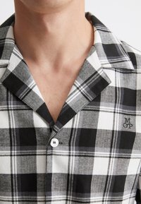 Musta-valkoinen ruudullinen paita, jossa on pehmeä kangas, klassinen kauluspaita, yksi valkoinen nappi ja brodeerattu logo rinnassa.