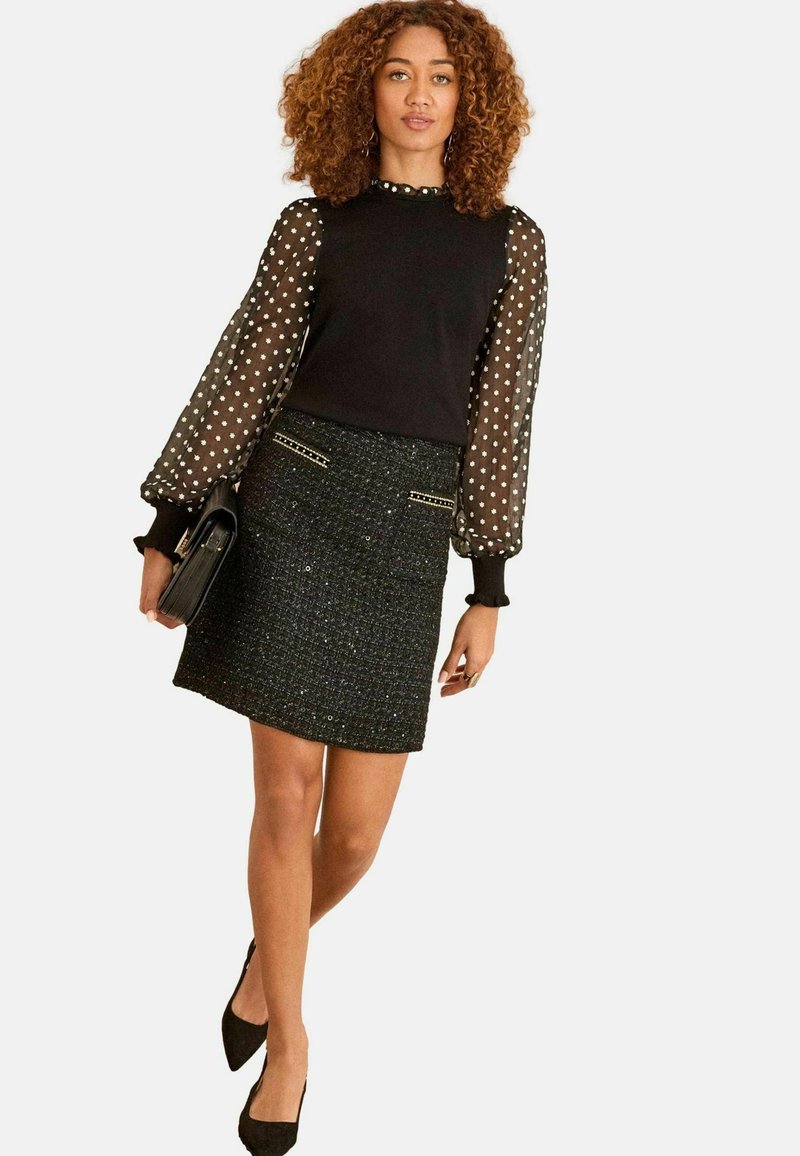 Črna bluza s prosojnimi rokavi s pikami, v kombinaciji s črno tweed krilom z zlatimi poudarki. Manekenka drži črno torbico in nosi črne pete.