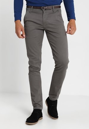 Hombre que lleva pantalones ajustados grises, un cinturón marrón, zapatos de ante negros y una camisa azul de manga larga, de pie contra un fondo blanco.