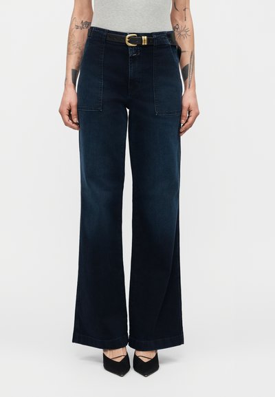 Personne portant un jean bleu foncé taille haute à jambes larges avec des poches avant plaquées, une ceinture noire avec une boucle dorée, et des chaussures noires pointues.