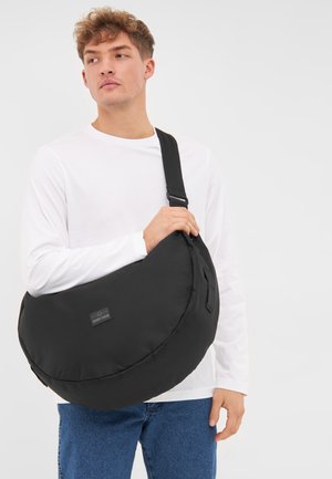 Borsa a spalla in tessuto nero con forma arrotondata, tracolla regolabile e un'etichetta con logo sul davanti. Indossata contro uno sfondo neutro.