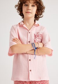 Roze shirt met korte mouwen van textuurstof, bruine knopen en rode kraagaccenten. Bevat borduurwerk "WILD SOUL" en "KIND HEART" op de borst.