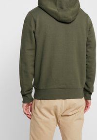 Felpa verde oliva con una superficie testurizzata, dotata di cappuccio, polsini e orlo a costine, indossata sopra pantaloni beige. Design semplice, senza motivi visibili.