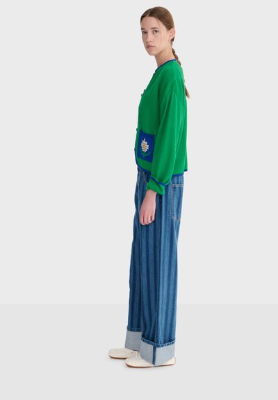 Femme en cardigan vert avec une poche fleurie, jean bleu large à revers, et chaussures blanches, debout de profil contre un fond clair.