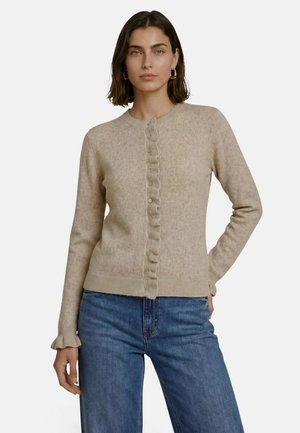 Kvinde med skulderlangt brunt hår iført beige cardigan med knapper og flæsekant samt blå jeans, står med den ene hånd i lommen.