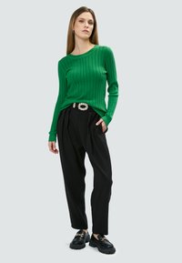 Maglione a coste verde abbinato a pantaloni a vita alta neri plissettati e mocassini neri con plateau, caratterizzati da una dettagli di catena dorata.