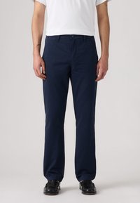 Homme portant un pantalon droit bleu marine, des chaussures habillées noires et une chemise blanche à manches courtes, debout devant un fond uni.