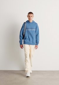 Felpa blu con logo "HUGO", pantaloni color crema, sneakers bianche e una borsa a tracolla grigia. Texture morbide e design casual.