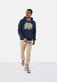 Marineblauwe hoodie met kleurrijke olifantprint en witte trekkoorden, gecombineerd met beige broek en grijze sneakers met zwarte accenten.