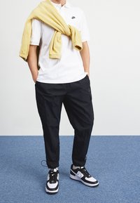 Valkoinen polo-paita Nike-logolla, mustat cargo-housut, joissa on tekstuurinen kangas, keltainen neule olalla, mustat ja valkoiset tennarit.