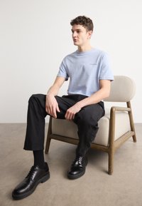 Lyseblå kortærmet t-shirt, sorte bukser og sorte lædersko. Model siddende på en beige mønstret stol foran en ensfarvet baggrund.