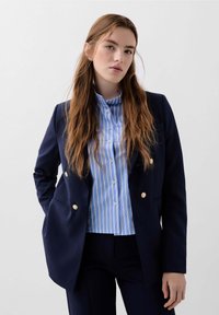 Námořnický blazer s akcenty zlatých knoflíků přes světlou modrou a bílou proužkovanou košili s knoflíky, v kombinaci s tmavými kalhotami. Hladká textura látky.