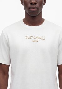 Черен човек, носещ бяла тениска със златен надпис "Just Cavalli Milano" на гърдите на обикновен фон.