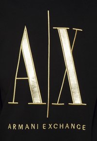 Sort stof med store, metalisk guld broderede bogstaver "A/X" og "ARMANI EXCHANGE" nedenfor i en elegant skrift.
