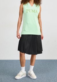 Ljusgrön ärmlös topp med vit kant och "PUMA"-logotyp, i kombination med en svart plisserad kjol och vita sneakers med grå detaljer.