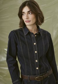 Robe chemise en denim bleu foncé avec des manches longues, accents en boutons dorés et coutures visibles. Une ceinture marron souligne la taille. Tissu texturé.