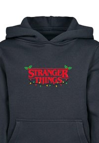 Tumši pelēks kapuce ar sarkanu "STRANGER THINGS" logo, kas rotāts ar zaļiem jāņogas akcentiem un krāsainām gaismām svētku dizainā.100% kokvilna.