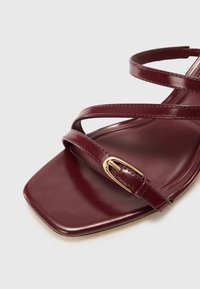 Burgundy högklackad sandal, tillverkad av glänsande läder. Har flera tunna remmar och en fyrkantig tådesign med ett guldspänne som accent.