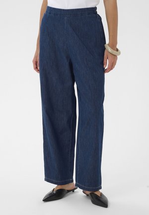 Sieviete valkā tumši zilas džinsa bikses ar augstu jostasvietu un platiem galiem, melnus lieljaudas mulekurpes ar smailu purngalu un bēša krāsas rokassprādzi uz labās rokas plaukstas.