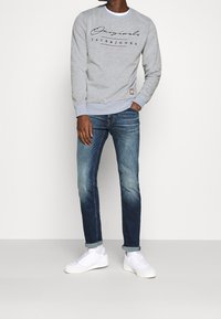 Felpa grigia con testo nero e rosso, jeans blu con effetto sfumato e sneakers bianche. La felpa presenta polsini e orlo a coste.